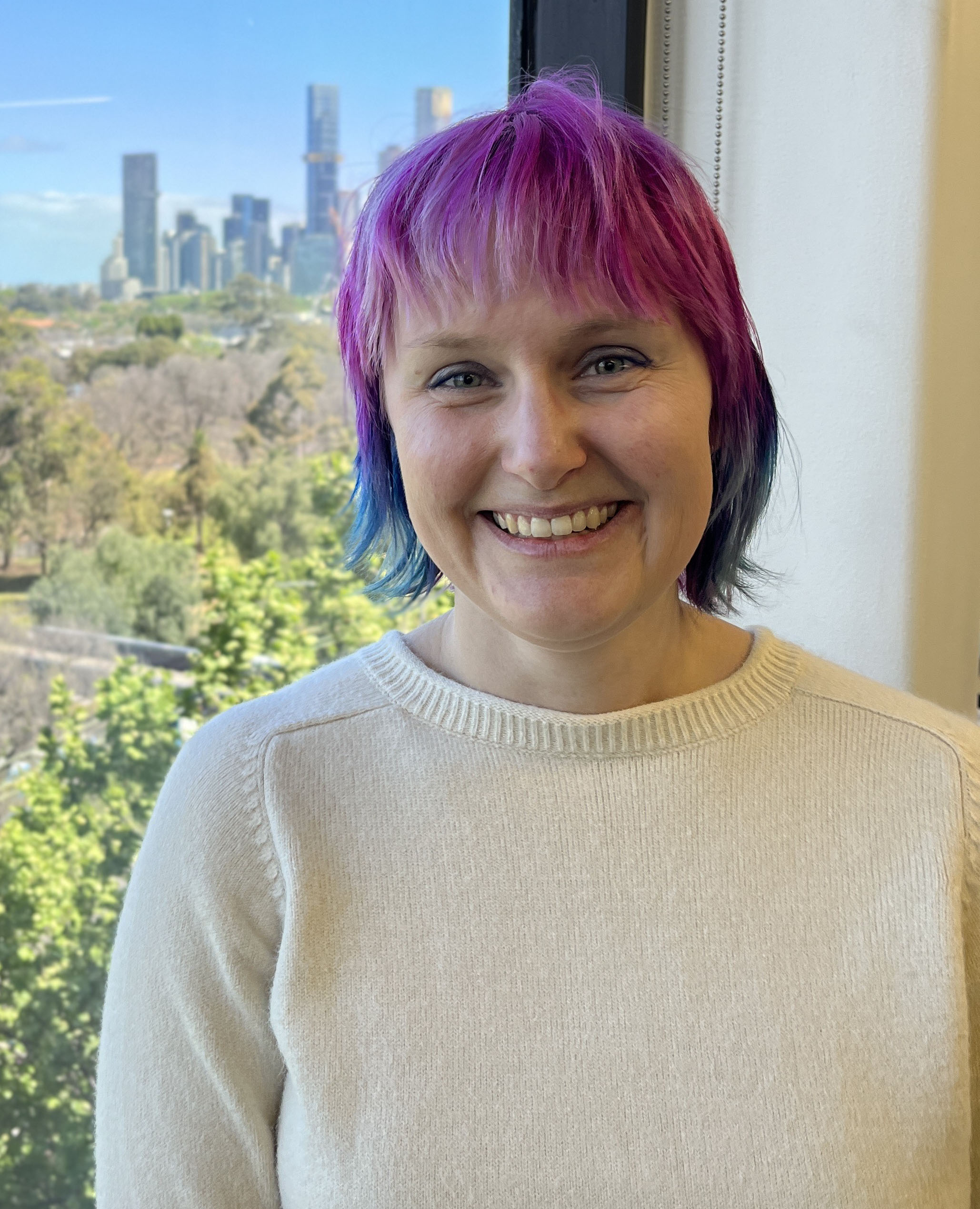 Danielle, a young white woman with rainbow hair, smiles for the camera. Her hair is pink on top and blue lower down and sits above her shoulders. She is wearing a white knit top and standing in front of window with a cityscape and garden behind her.