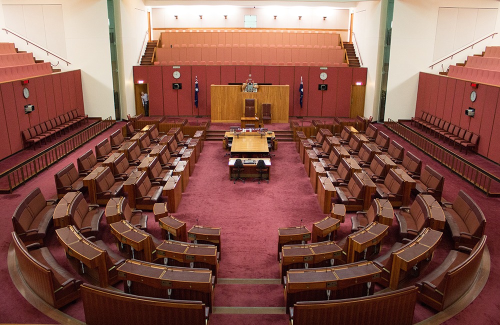 We are looking down into the Australian senate. It is a red room with empty red bench chairs around it and wood panelling on the walls.