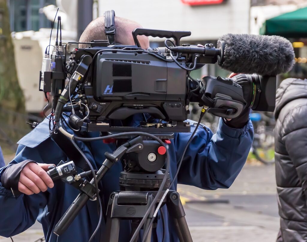 A person standing behind a large film camera, with a fluffy microphone on the front. The person's face is hidden by the camera.