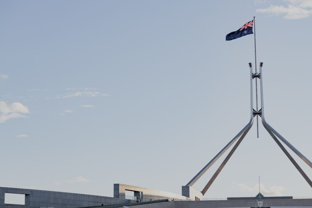 The flag on top of Parliament House, pictured from a distance, against a grey sky. The flag is on a big triangular flagpole.