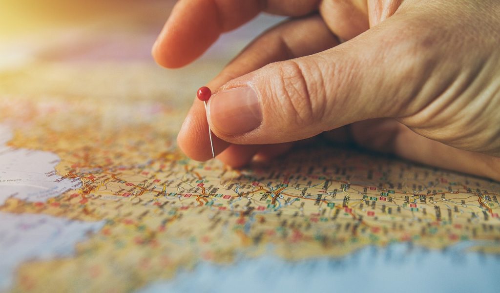 A hand holds a red pin just above a brown-toned map. The map is out of focus so we can't see the place it shows.