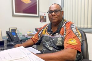 Ross Andrews, a Kuku Yalanji and Djungan man, sits at a desk in an office. He has very short hair and is wearing a bright top with black and orange colours on it.