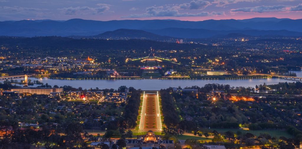 A view of Canberra at night, looking from the War Memorial down to Parliament House, over Lake Burley Griffin. The buildings are lit up and the road between them is lit up gold.