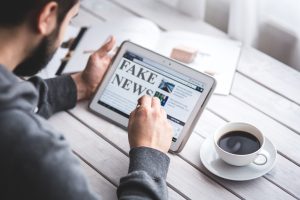 A man sits at a wooden table with a black coffee in a white mug beside him. He has a beard and is wearing a grey top. We are looking down from behind at the tablet in his hands, which says 'fake news'.