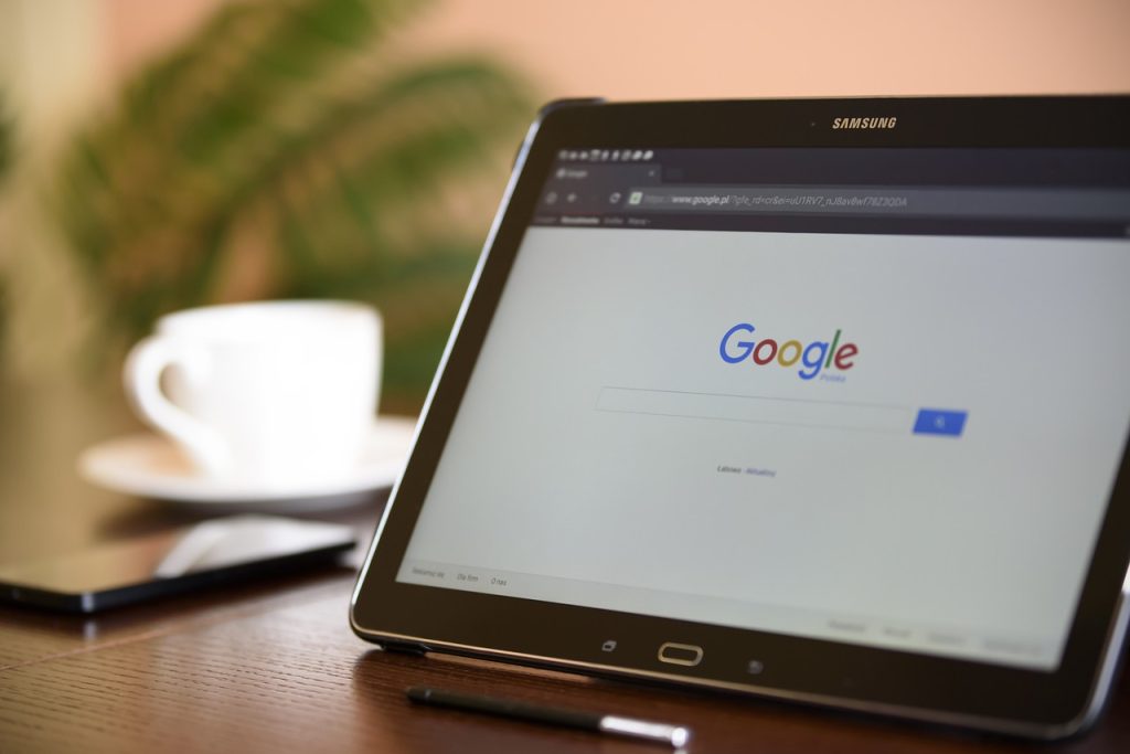 In the background is a white mug on a desk. In the foreground, a computer tablet sits open with the homepage of Google displayed.