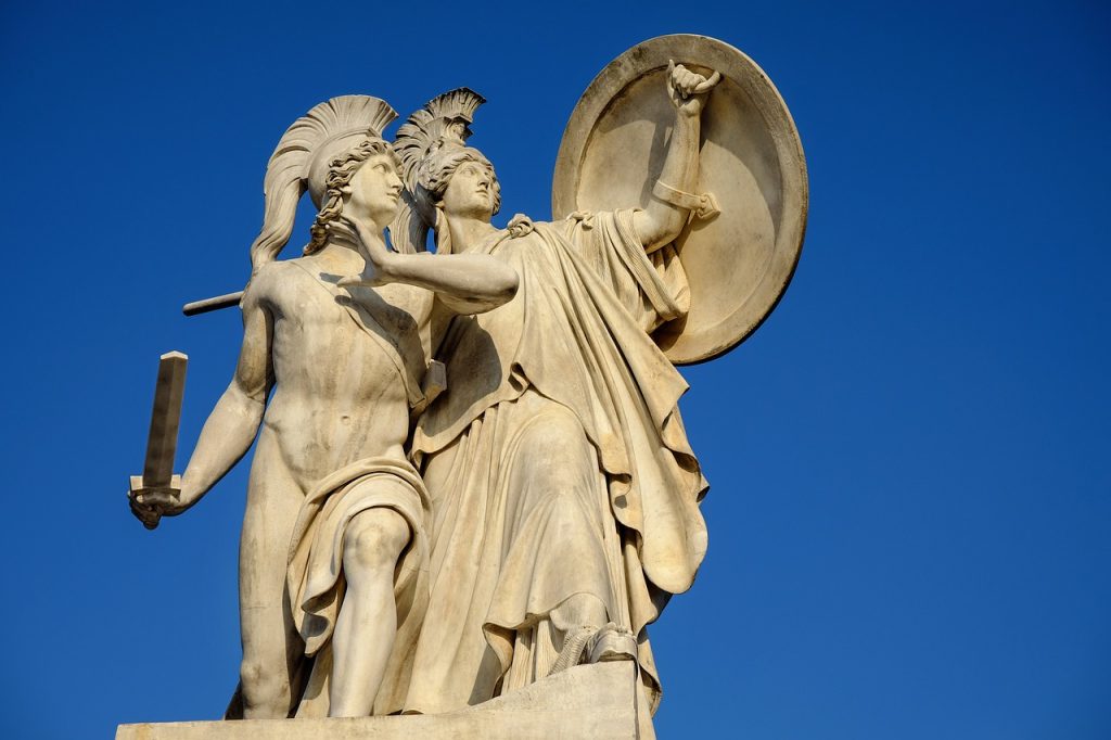 A statue/monument against a blue sky. The statue is a pale stone and depicts a man holding a sword, naked, and another person in a robe holding a shield above them both. They are both wearing helmets and look to be ancient Greek.