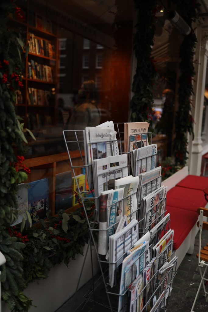 A stand of newspapers outside a shop. The shop is in darkness and the papers are in the foreground and lit up.