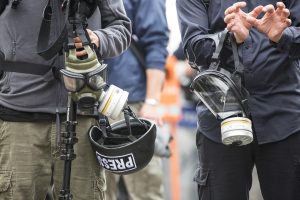 Members of the press. One person is holding a gas mask and a helmet that says 'press'. The other person is carrying a gas mask too and is holding their hands out like they are about to be arrested. We can't see their faces.