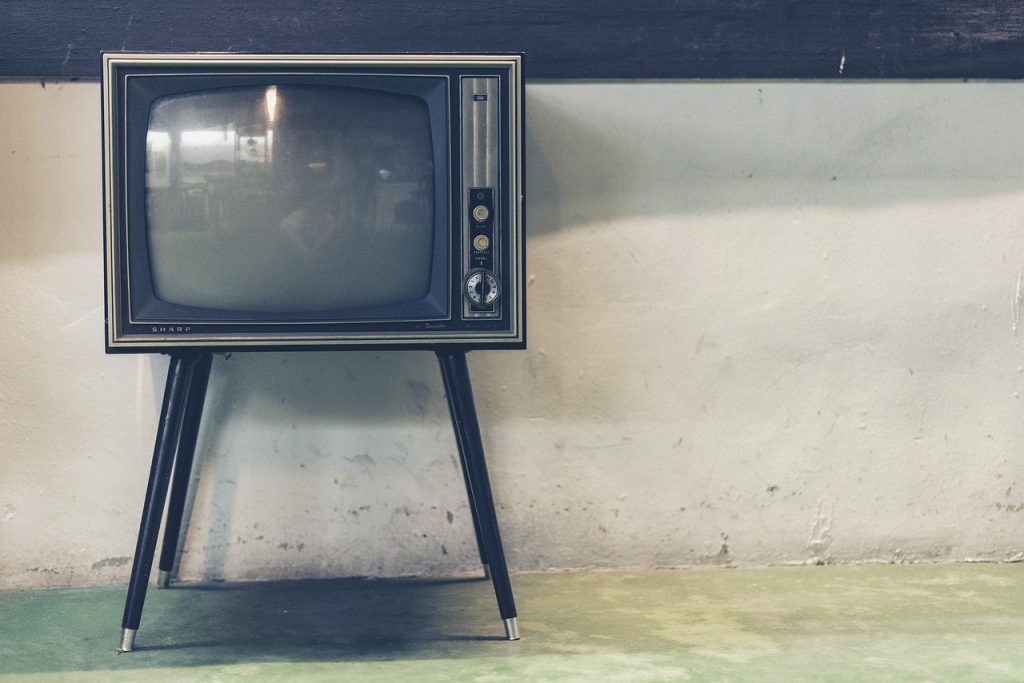 An old-fashioned black television on four black outward-angled legs, sitting in front of a wall.