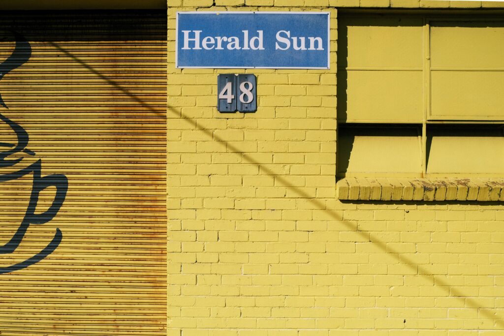 A yellow painted brick wall with a blue sign on it that says Herald Sun, and the house number 48 beneath it.
