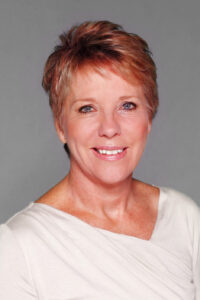 Anita Jacoby smiles at the camera. She is a middle-aged woman, with close cropped hair in an orange-blonde tone and wearing a white v-neck shirt. We only see her from the shoulders up.