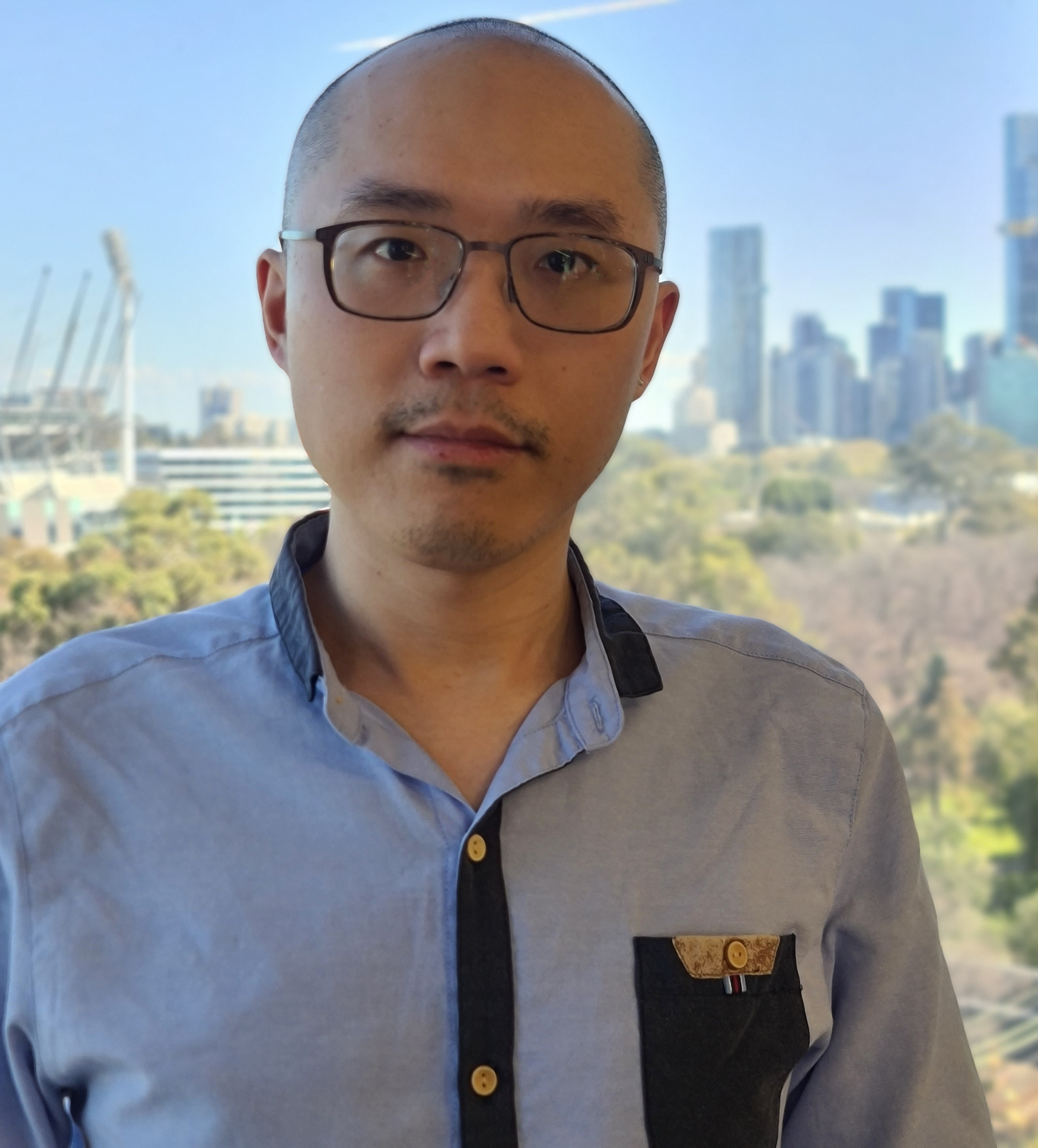 Erik Wu, a young Asian man, stands in front of a window with a cityscape and gardens behind him. He has no hair, and wears black rimmed glasses. He is wearing a blue button up shirt with black details around the buttons, pocket and collar.