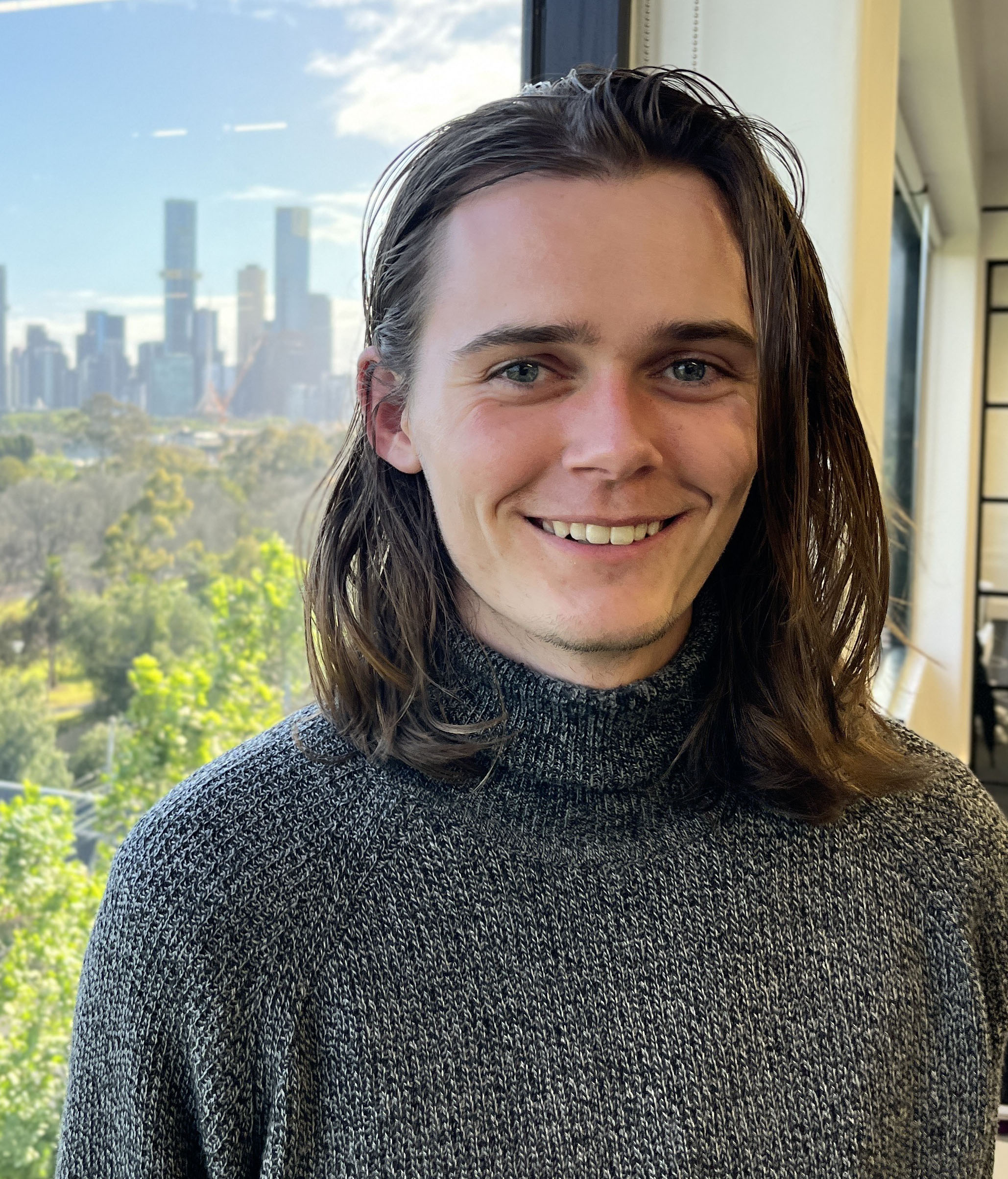 Jerome Des Preaux, a young man, smiles at the camera. He has shoulder length brown hair and is wearing a dark grey turtleneck jumper. He is standing in front of a window with a city scape and gardens behind him.