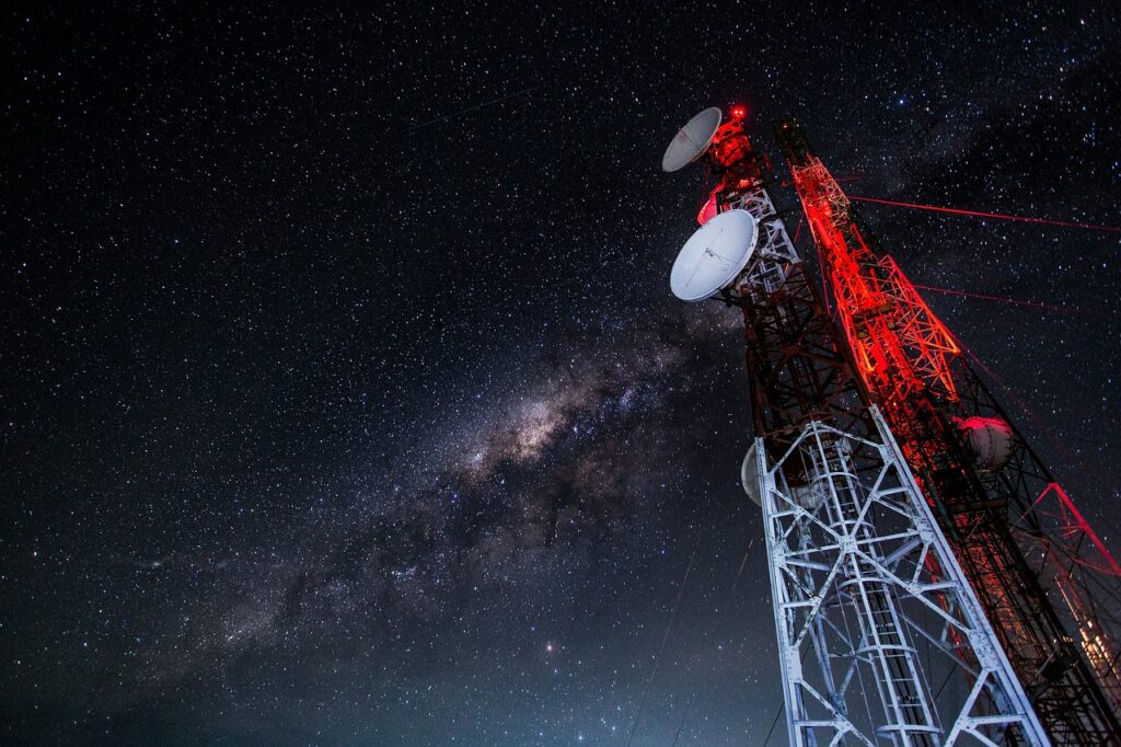 A broadcast/radio signal tower against a dark, starry night sky. The tower is lit in red at the top and silver at the bottom.