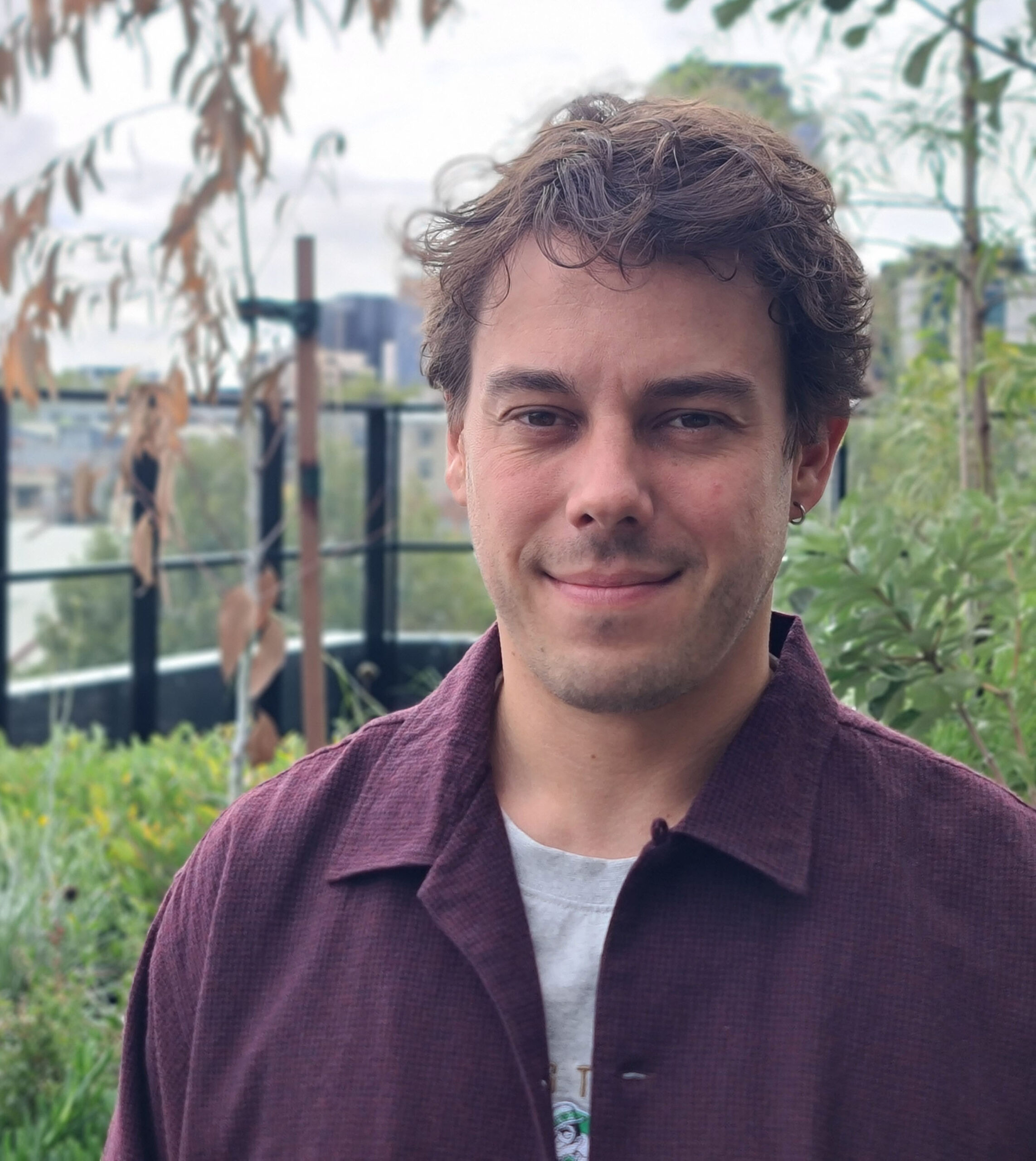 Jacob, a white man, stands before the camera. He is standing in a garden and wearing a maroon button up shirt over a grey tshirt. He has brown hair that is slightly curly.